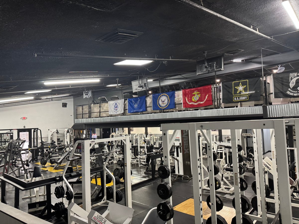 Military-themed gym with exercise machines, military flags on upper wall, and yellow and black equipment visible throughout the facility
