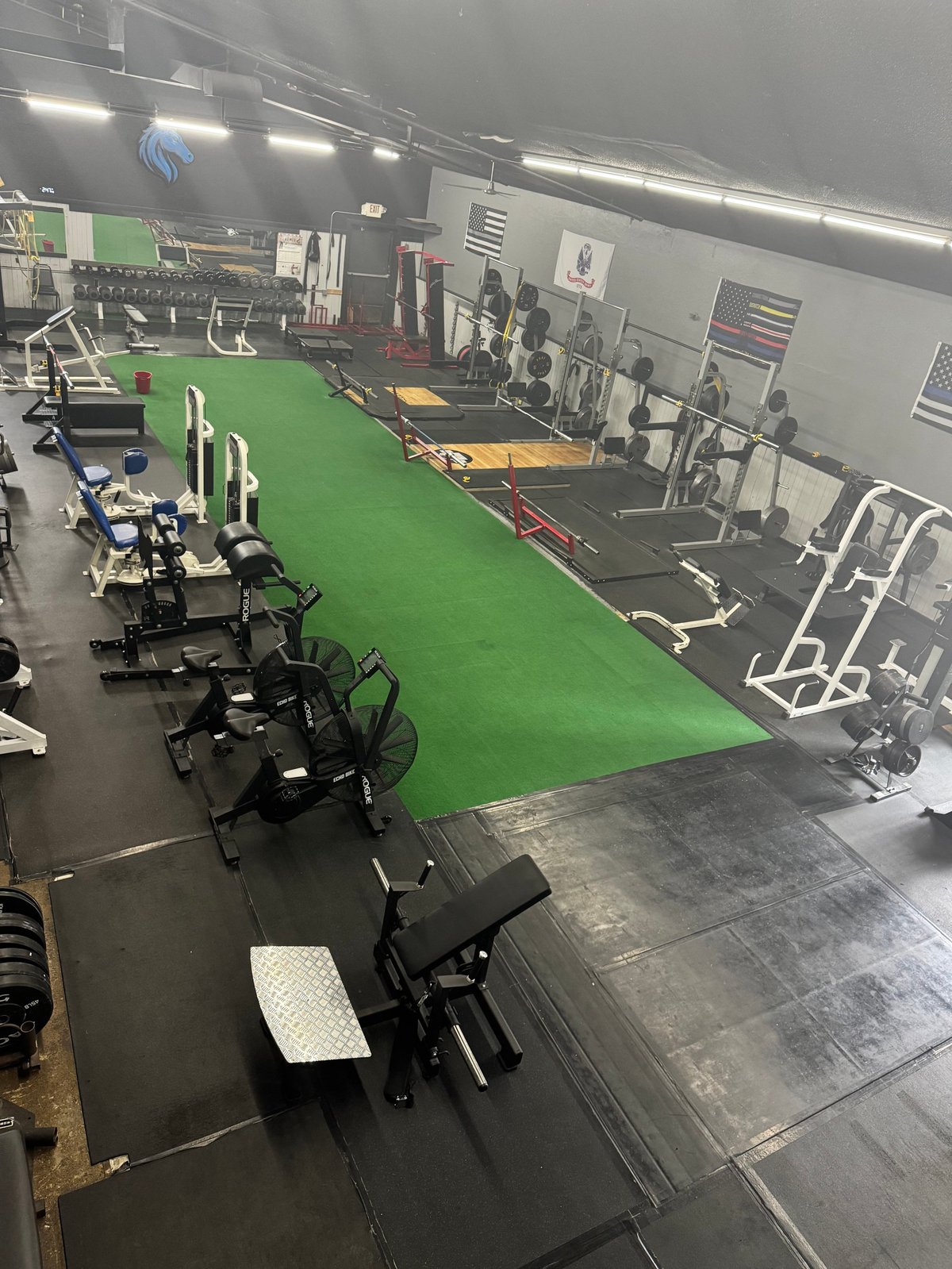 Indoor gym with bright green turf running track, stationary bikes on left, weightlifting equipment and weights rack on right, concrete and rubber flooring, fluorescent lighting